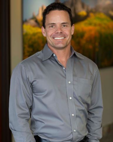 Professional man in gray button-up shirt smiling at camera in modern office setting.