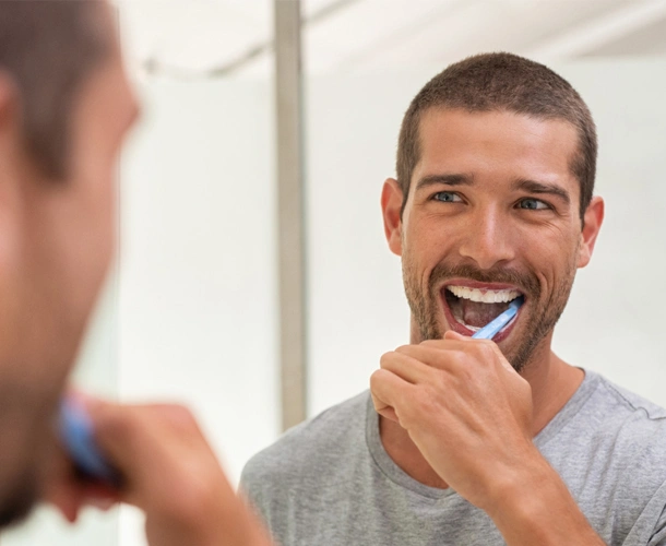 Man brushing teeth in bathroom mirror, smiling at reflection.