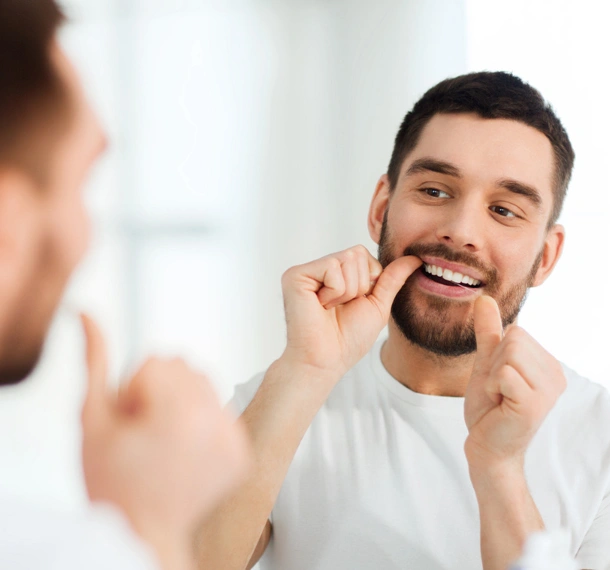 Man flossing teeth while looking in bathroom mirror, smiling.