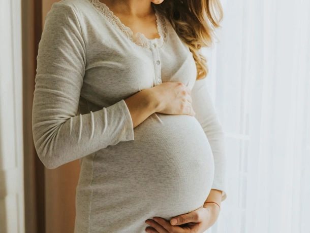 Pregnant woman in gray clothing holding her belly against a white wall indoors.