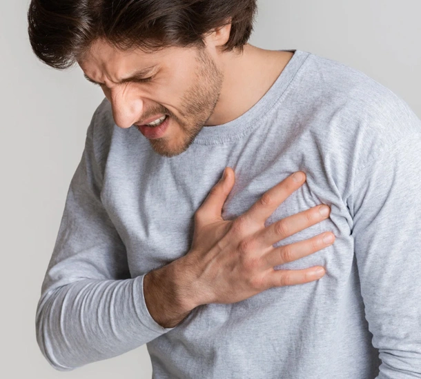 Man in gray shirt holding chest, appearing to experience discomfort or pain indoors.