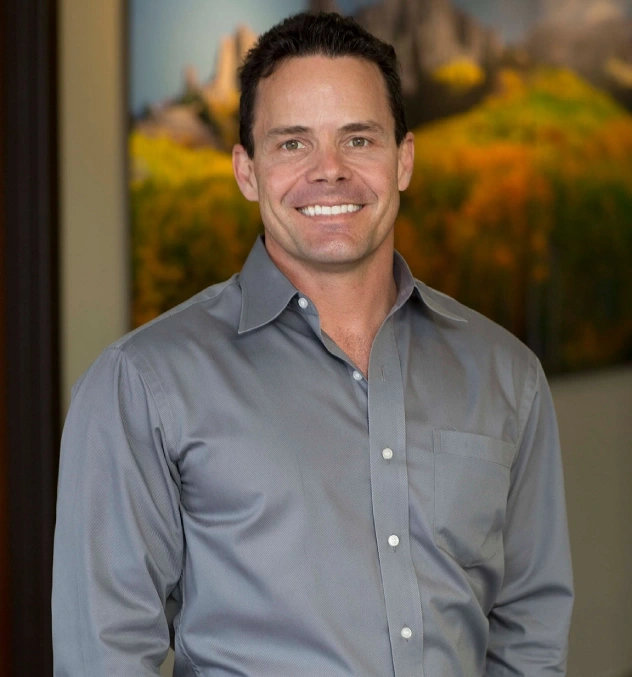 Professional headshot of smiling man in gray button-up shirt against blurred office background.