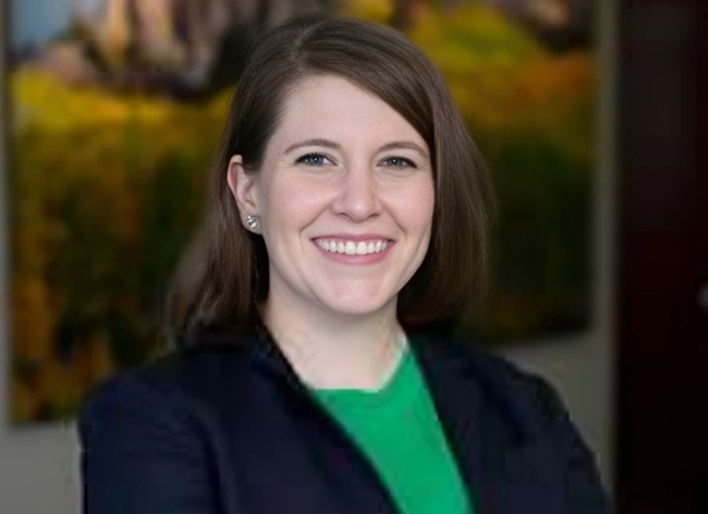Professional headshot of smiling woman in green shirt and dark blazer at dental office.